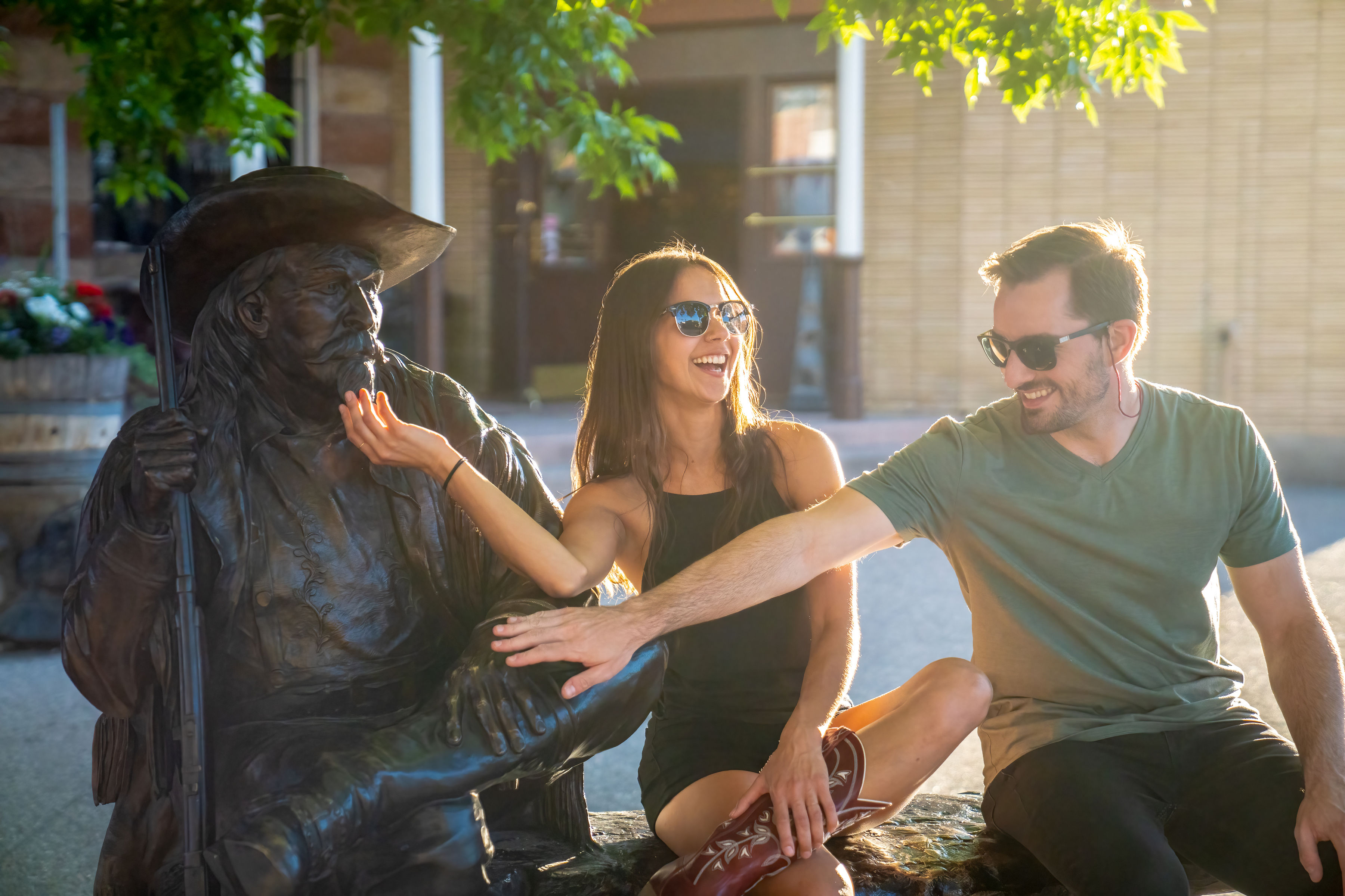 A man and woman sit next to the statue of Buffalo Bill Cody and playfully tug his beard