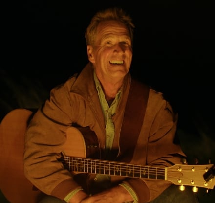 Dan Miller sitting with his guitar in front of a fire
