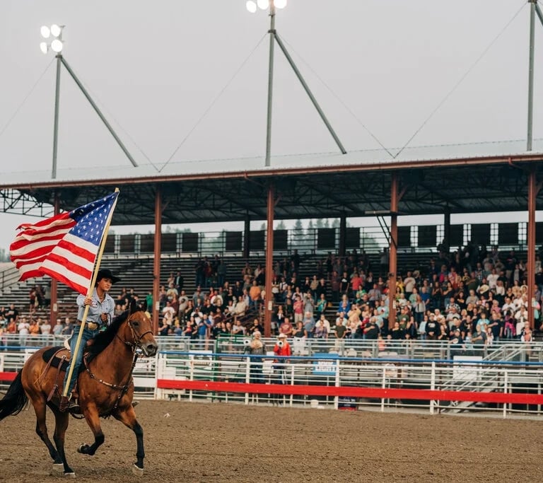 A cowboy on a horse holds an American flag high as he rides before a lively crowd