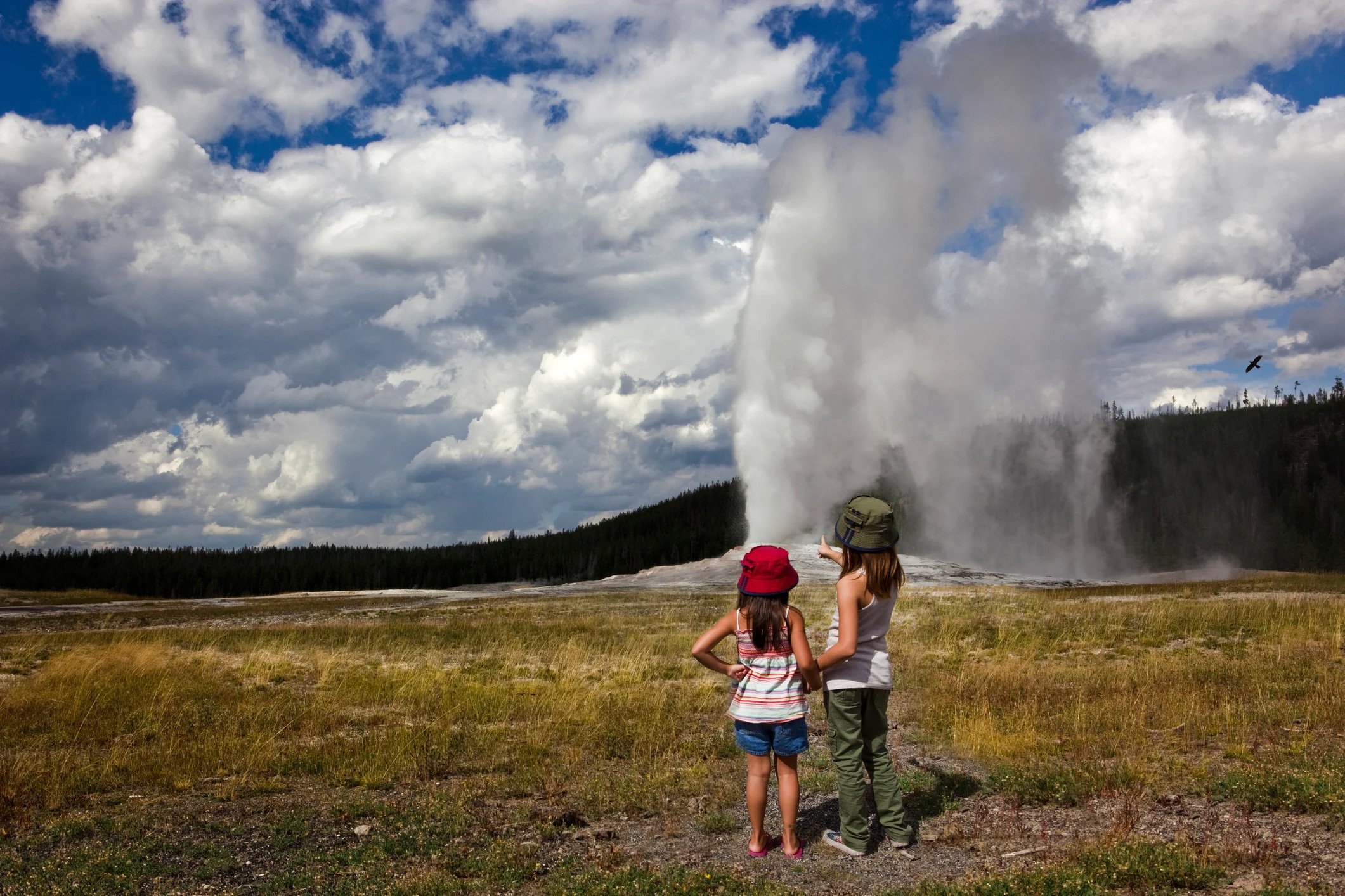 Two kids gaze in awe at a geyser erupting in Yellowstone National Park, surrounded by stunning natural beauty