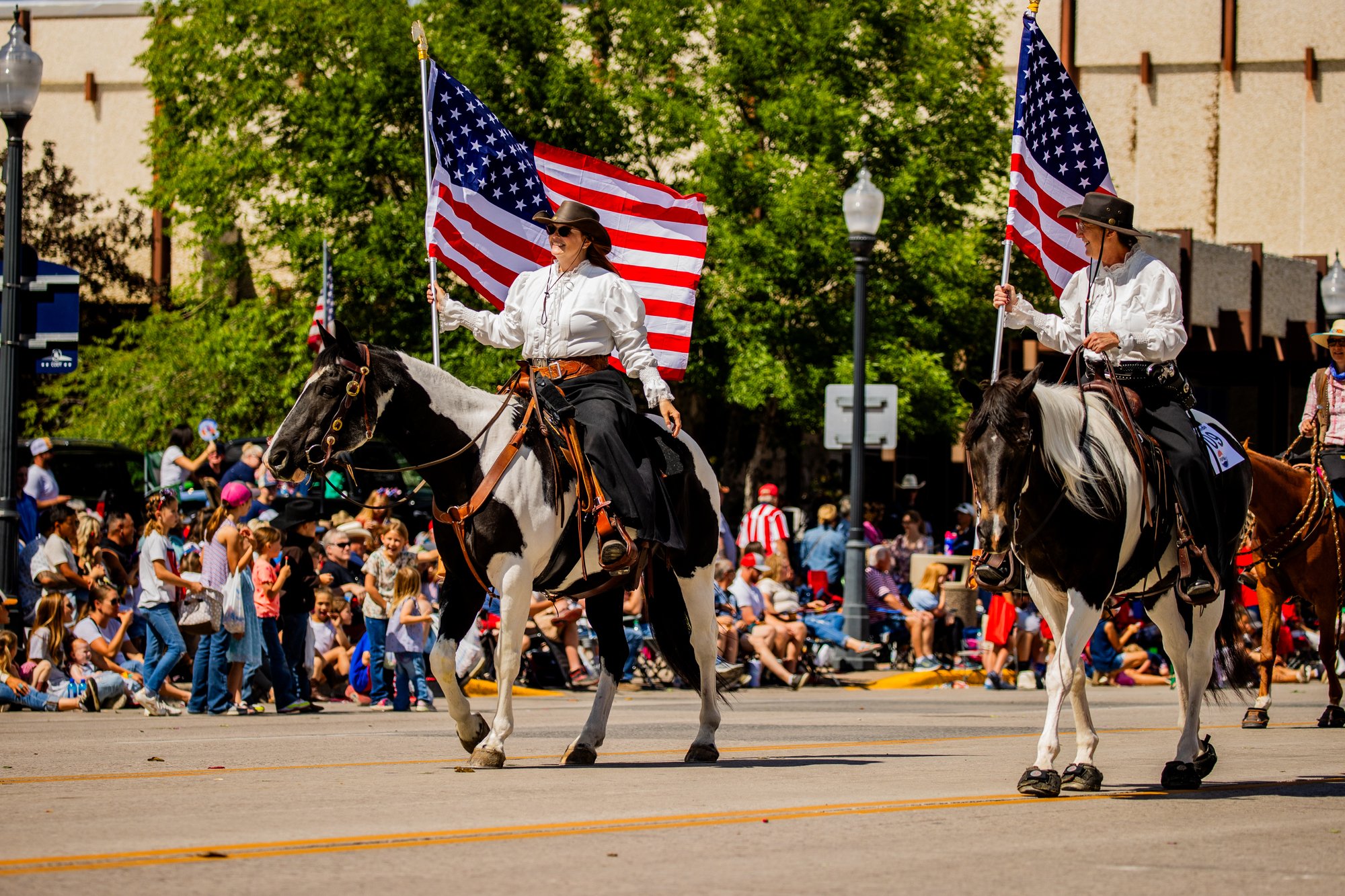 Two people ride horses while carrying American flags at the July 4th Parade in Cody Yellowstone
