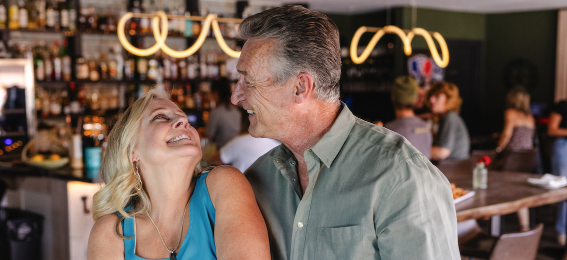 A smiling older couple standing close together in a cozy bar or restaurant, looking into each other’s eyes and holding hands, with warm lights and other patrons blurred in the background.