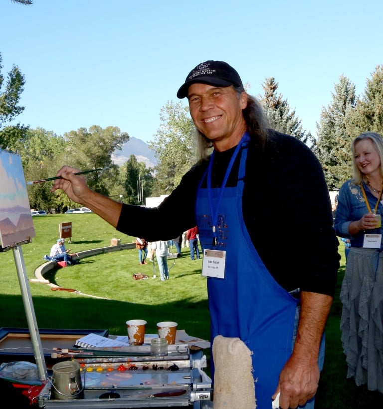 A man wearing a blue apron painting a landscape painting in the park
