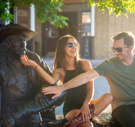 A man and woman sit next to the statue of Buffalo Bill Cody and playfully tug his beard