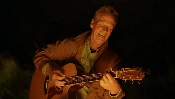 Dan Miller is sitting next to a campfire and strumming an acoustic guitar and singing.