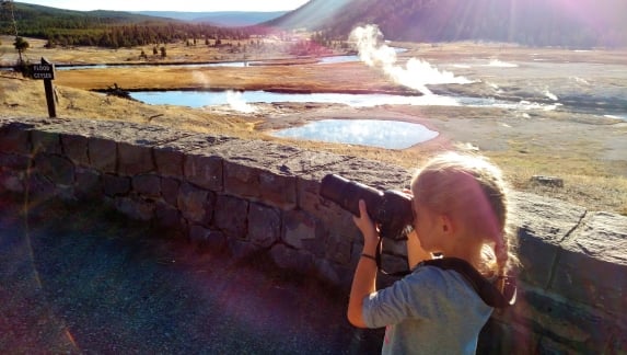 A blonde girl in a pony tail taking a photo with a geyser in the background at Yellowstone National Park.